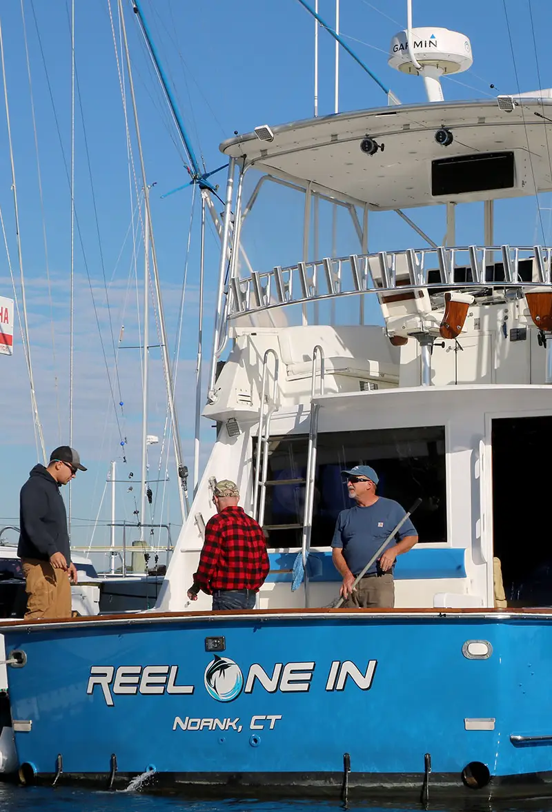 trip-2 about section image Three people are on a fishing boat named "Reel One In," docked in a marina with clear blue skies and rigging in the background.